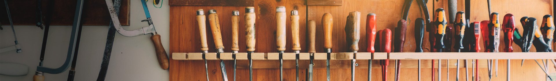 Eine Wand mit verschiedenen Handwerkzeugen, die ordentlich an einem Holzregal aufgehängt sind. Die Werkzeuge umfassen Holzgriffmeißel, Schraubendreher und eine Handsäge. Der Hintergrund ist aus Holz und die Werkzeuge sind in verschiedenen Farben und Größen